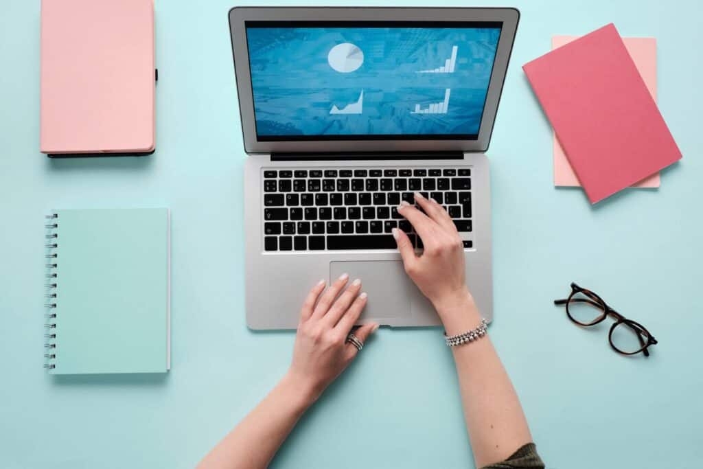 View of a digital marketing professional's hands typing on a silver laptop on a blue desk with pink notebooks.